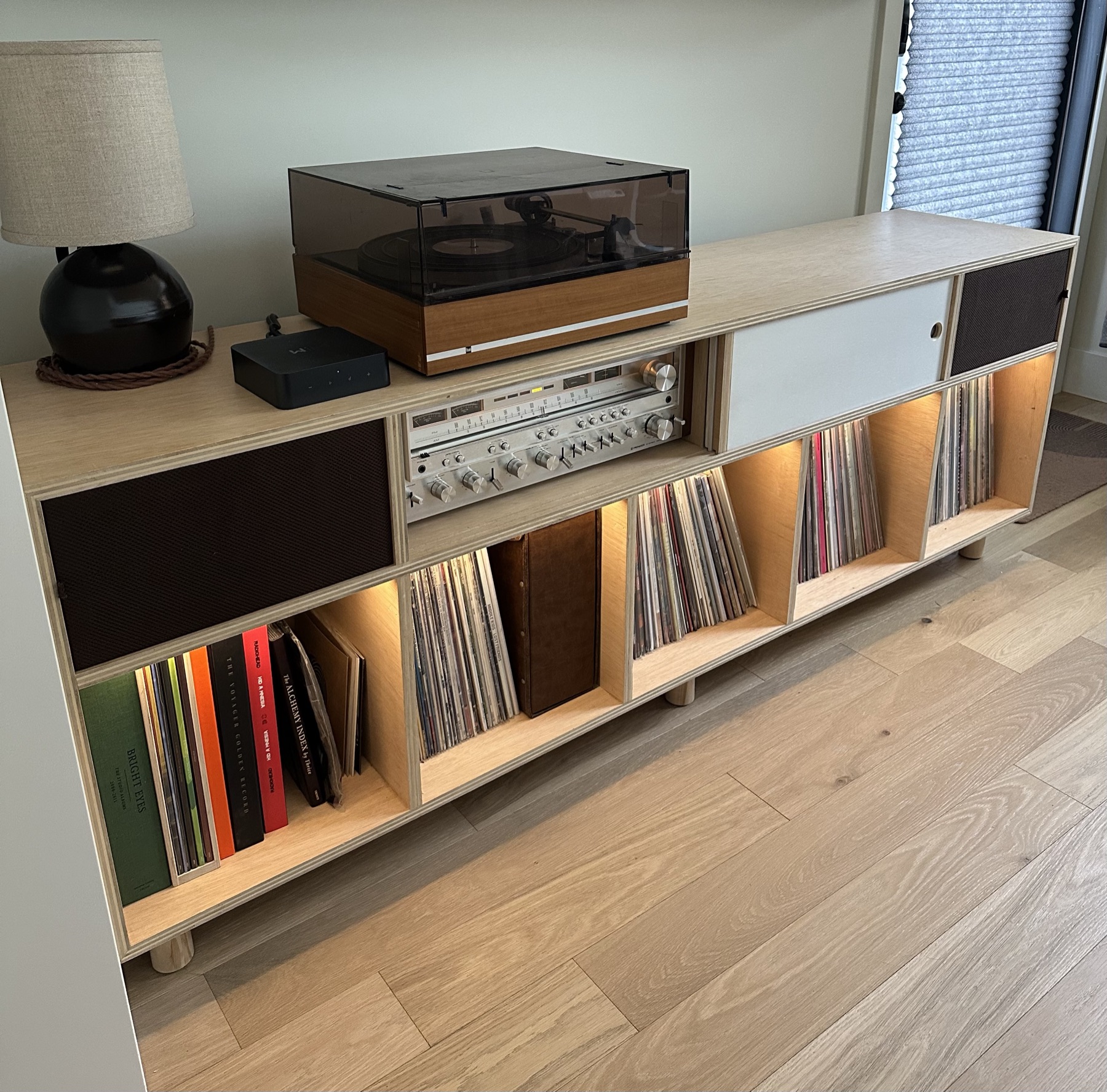 Angled view of the plywood record cabinet with a turntable on top, a stereo receiver behind an open sliding door, and lit shelves full of records