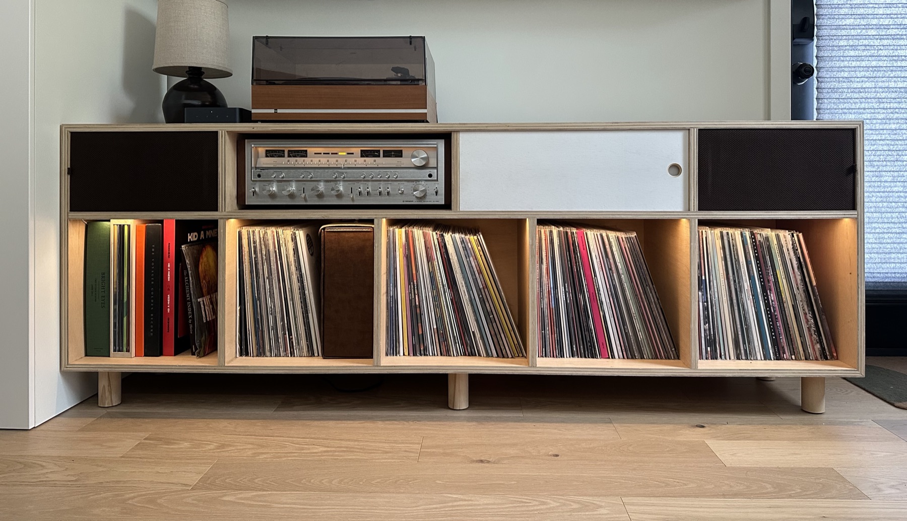 Front view of the record cabinet with its sliding door open to show the receiver, speakers behind dark panels at each end, and records below
