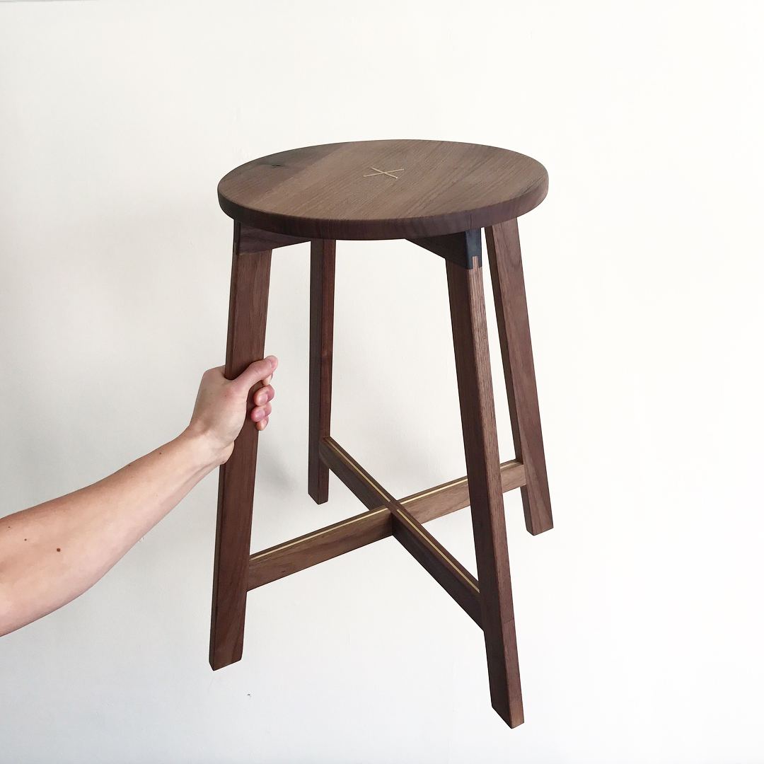 A hand holding up a tall walnut stool with a round seat, splayed legs and crossed stretchers