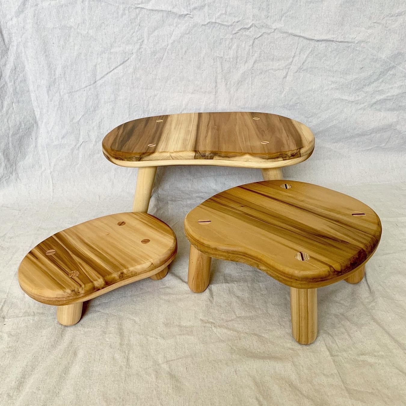 A set of three low poplar stools with rounded, bean-shaped seats on a linen backdrop